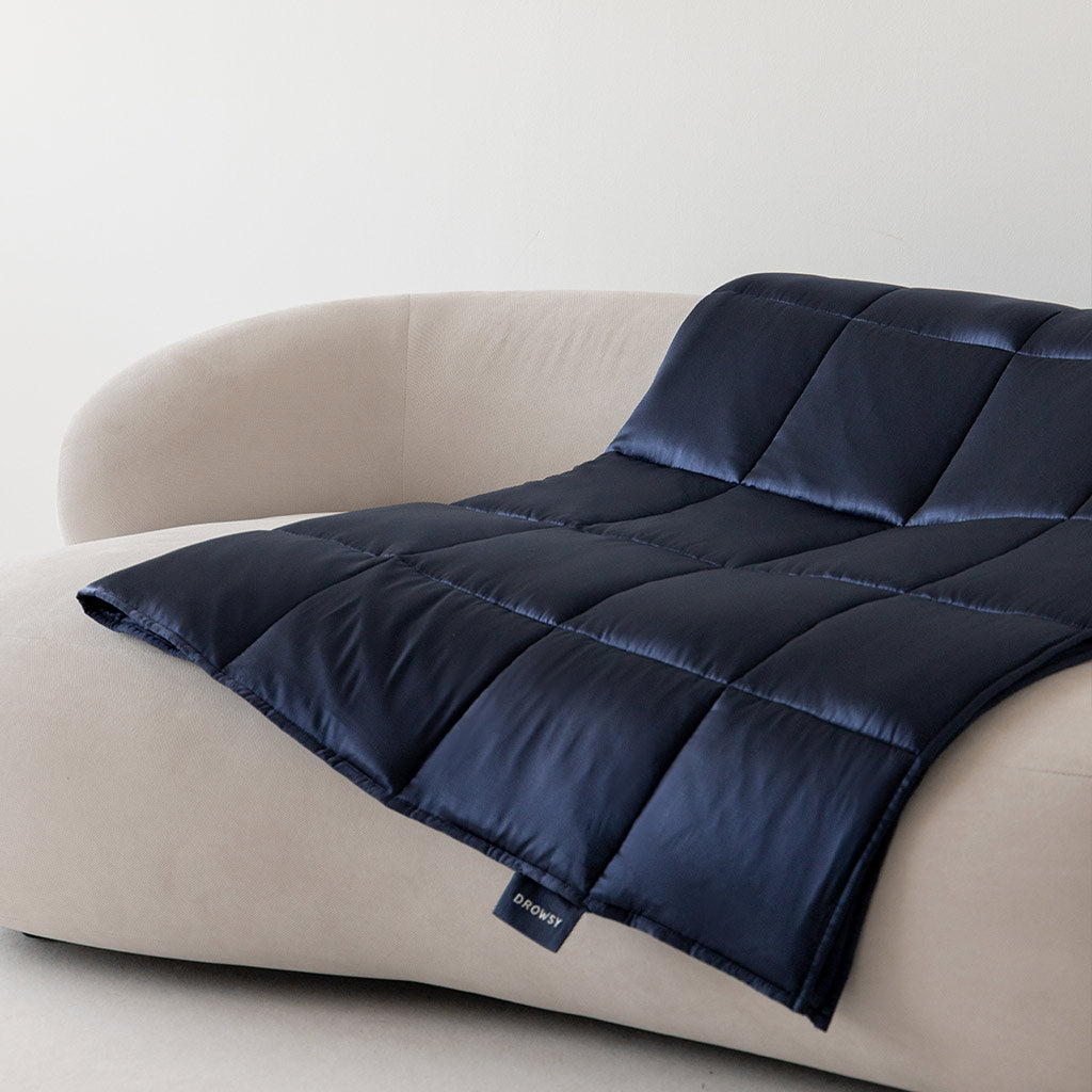 The Midnight Blue Silk Weighted Blanket is neatly draped over a light beige, curved sofa against a plain white wall. A small fabric tag on the edge hints at deep relaxation and comfort.