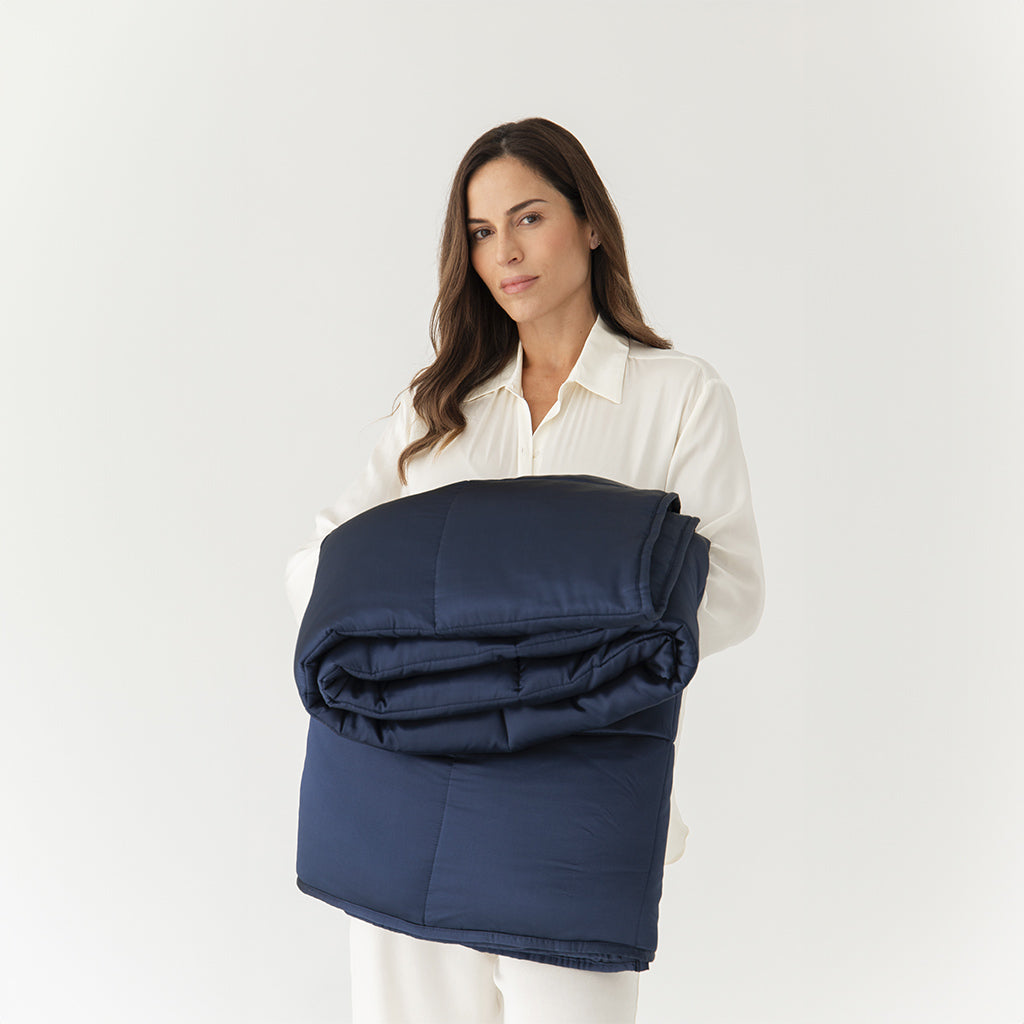 A woman with long brown hair in a white outfit stands against a plain white background, holding a neatly folded Midnight Blue Silk Weighted Blanket in her arms.