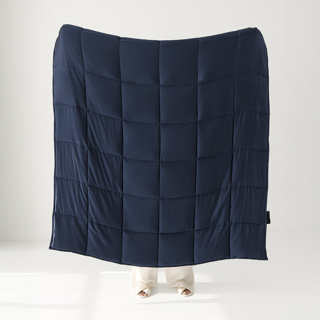 A person holds up the Midnight Blue Silk Weighted Blanket, its large quilted silk design covering most of their body and revealing only their feet, set against a plain, light background that evokes deep relaxation.