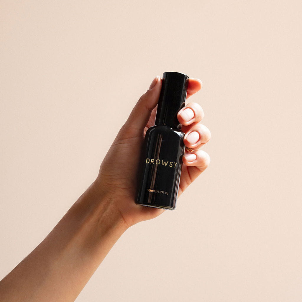 A hand holds a black bottle labeled Sleep S.O.S Pillow Spray, a natural sleep aid with a sleek, minimalist design and gold lettering, set against a neutral beige background—perfect for your nightly ritual.