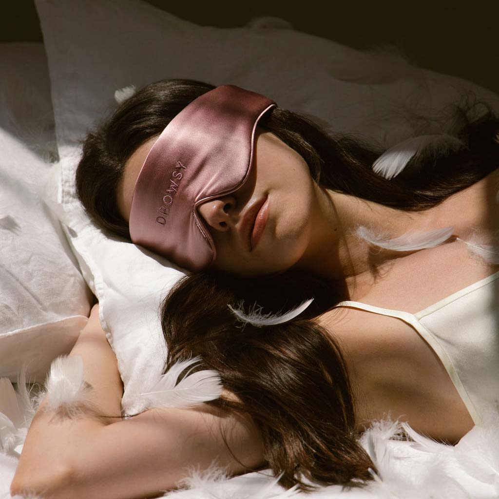 A woman rests on a bed with white sheets and feathers, wearing a silky pink sleep mask and a white camisole. She is lying on her back with one arm under her head.
