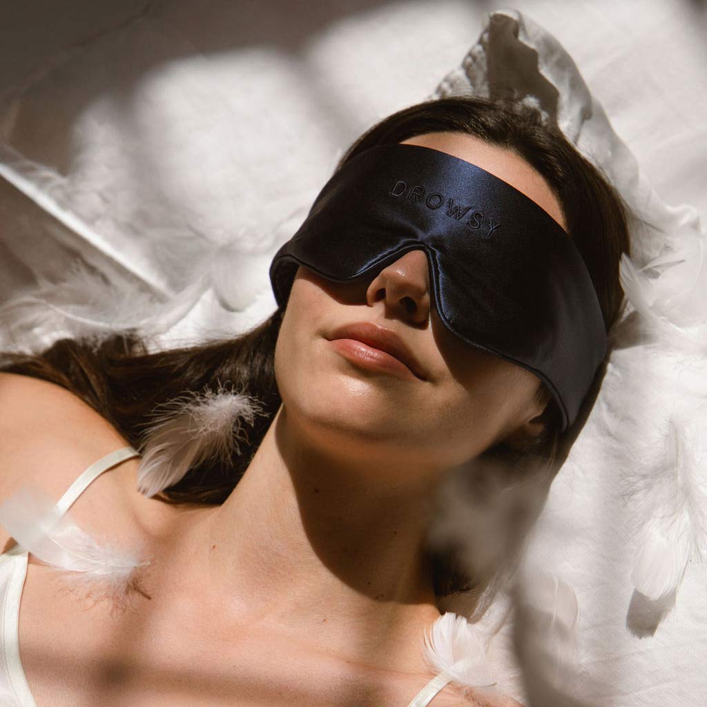 A woman lies on white sheets wearing a dark satin sleep mask with DROWSY embroidered on it. Soft feathers are scattered around her, and sunlight creates patterns across the bed.