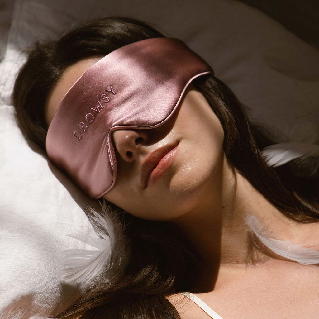 A woman lies on her back in bed, wearing a shiny pink sleep mask with the word DROWSY embroidered on it. She has long dark hair and is resting on a white pillow with a feather nearby.