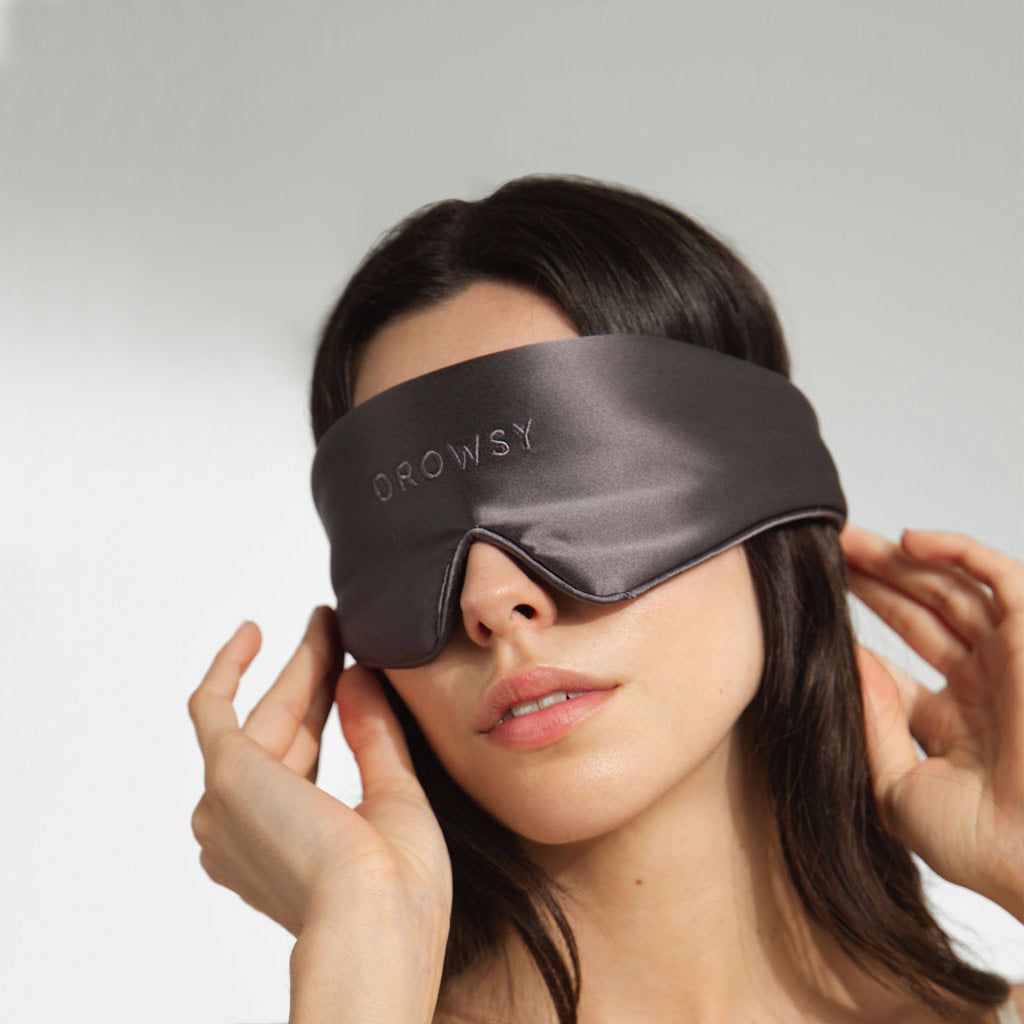 A woman wears a dark gray silk sleep mask with the word DROWSY embroidered on it, gently adjusting the mask with her hands against a light background.