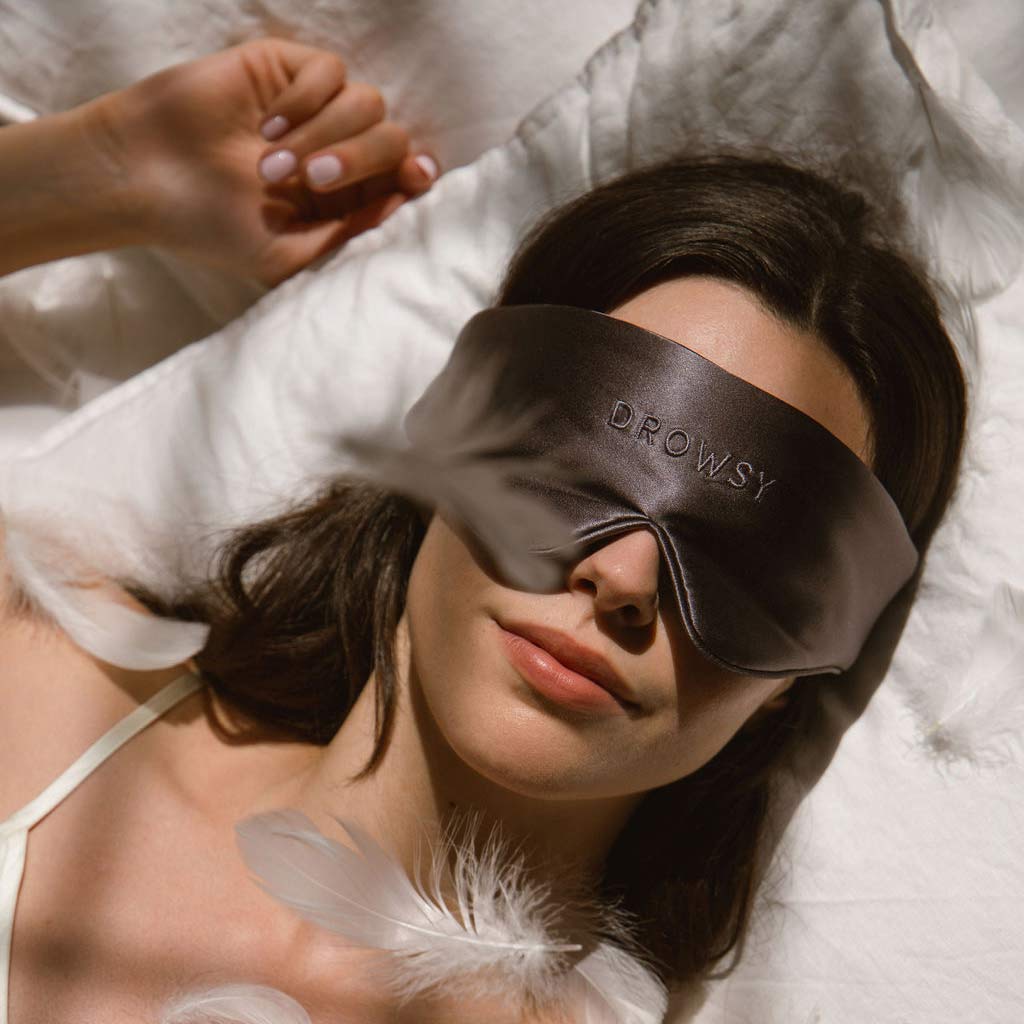 A woman lies on a white pillow wearing a satin sleep mask that says DROWSY. She is surrounded by white feathers, and her hand is resting next to her head. Soft light creates a calm, restful atmosphere.