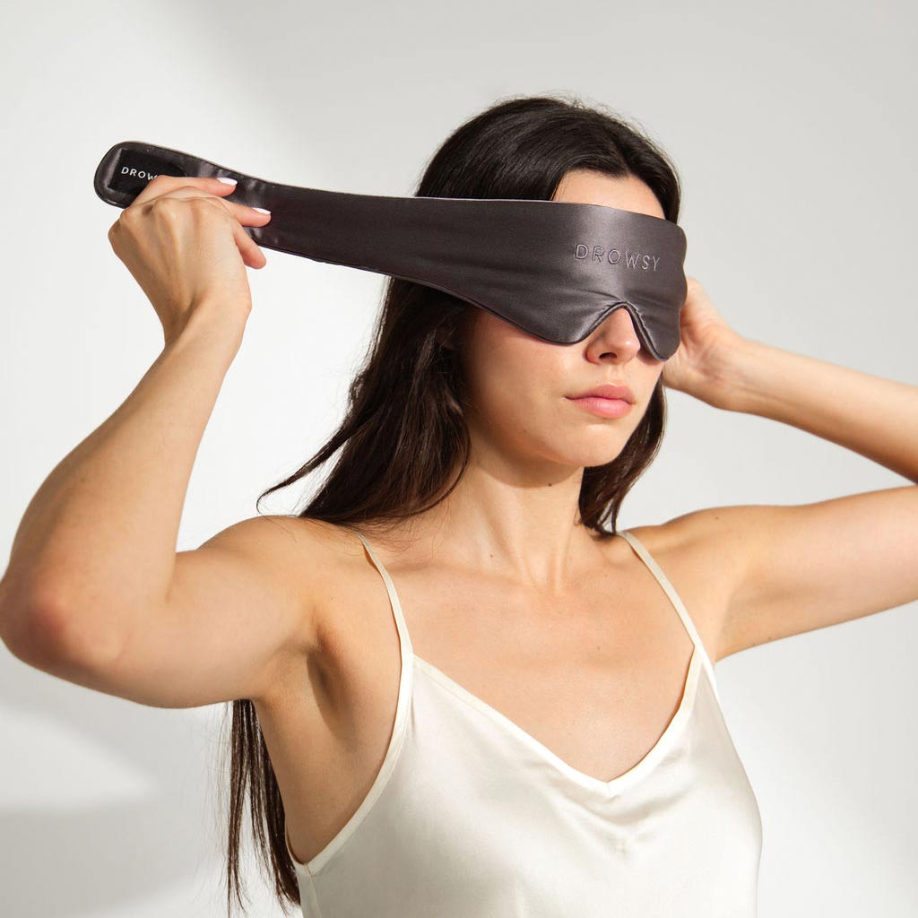 A woman in a white satin camisole puts on a Moonlight Shadow Featherweight Sleep Mask, pulling the strap over her head.
