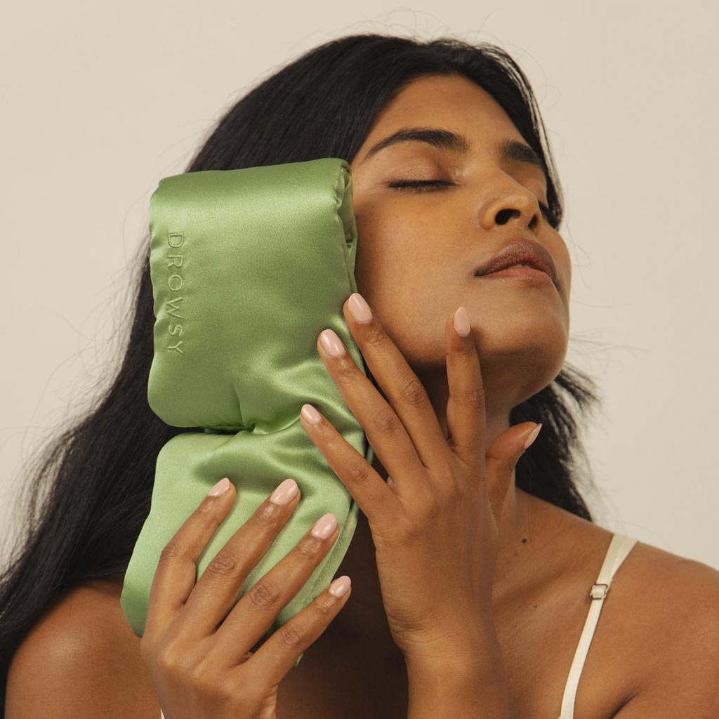 A woman with long dark hair holds a Pistachio Green silk eye mask to her face, eyes closed and head tilted back, appearing relaxed and ready for beauty sleep.