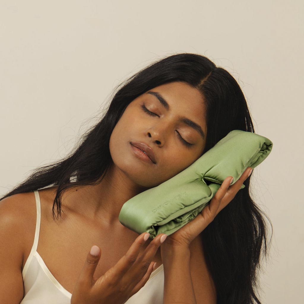 A woman with long dark hair and closed eyes gently rests her cheek against a soft, light green satin pillow, appearing calm and relaxed, against a plain light background.