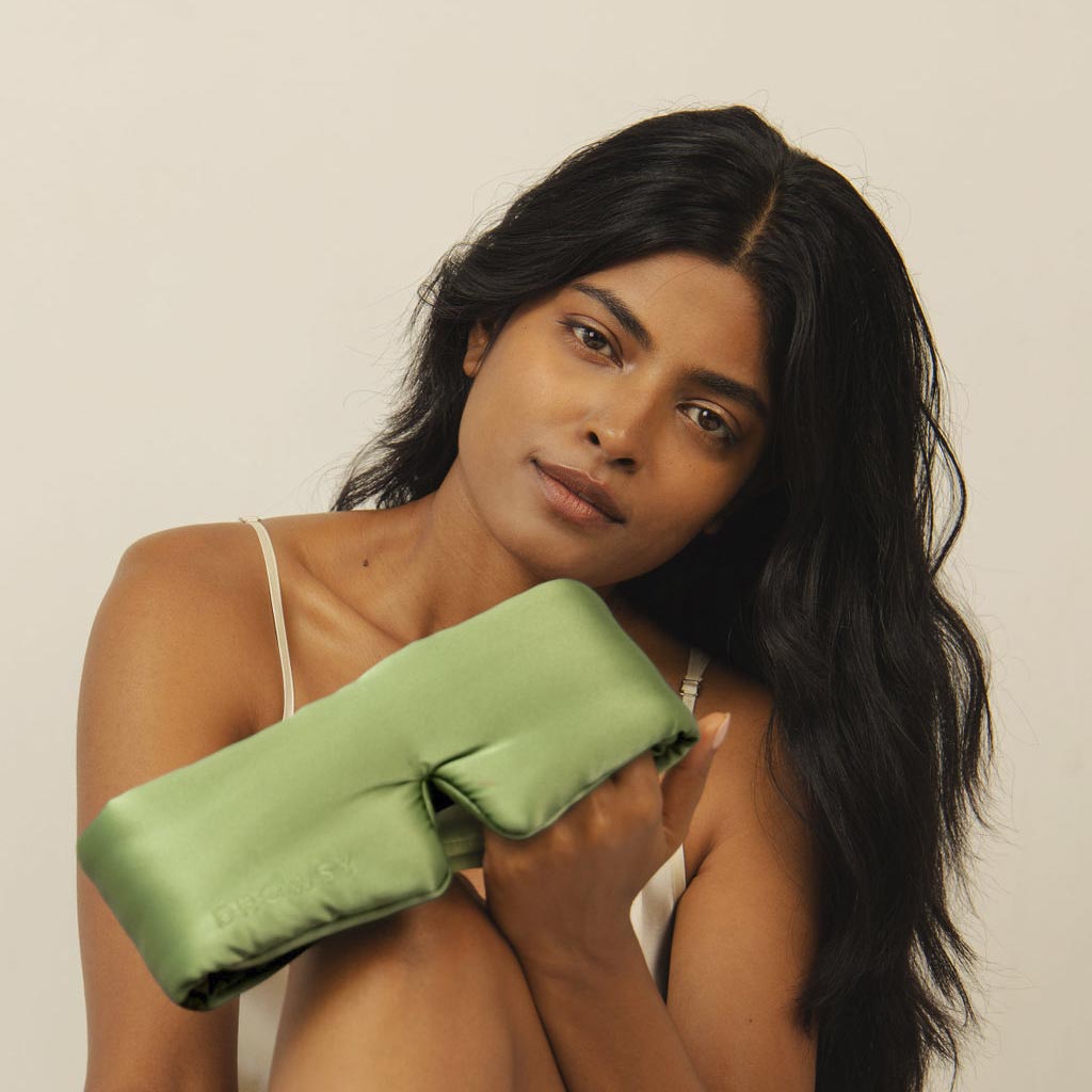 A woman with long, dark hair sits against a neutral background, wearing a white camisole and holding the Pistachio Green satin-covered, T-shaped blackout sleep mask, gazing softly at the camera.
