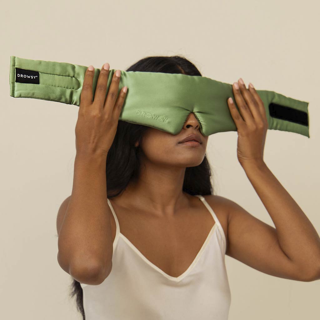 A woman in a white camisole holds the Pistachio Green silk eye mask over her eyes, embracing beauty sleep against a plain beige background.