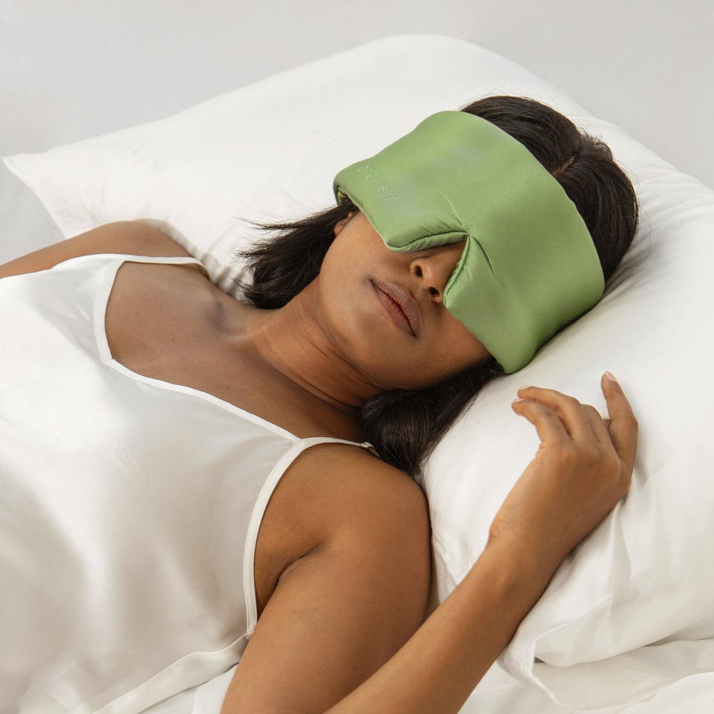 A woman in a white satin sleepwear lies on a white pillow and bed, wearing a large green sleep mask that covers her eyes and part of her nose, appearing relaxed and restful.