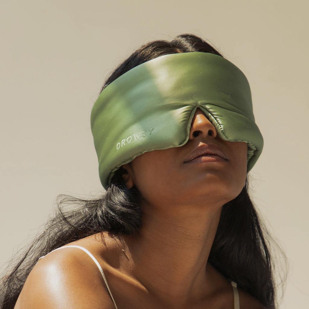 A woman with long dark hair wears a large, padded green sleep mask labeled Drowsy while looking upward. She is dressed in a light-colored sleeveless top against a neutral background.