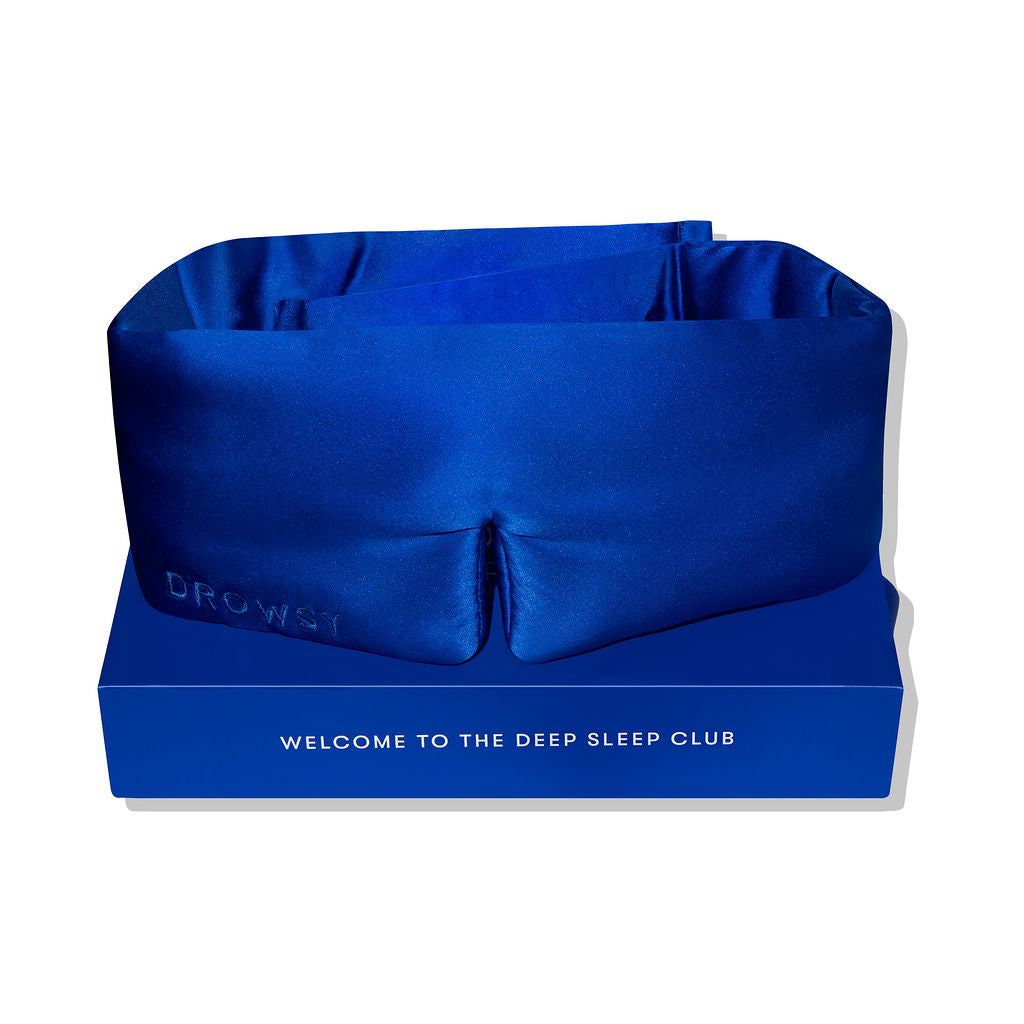 The Royal Blue silk eye mask rests on a matching box reading WELCOME TO THE DEEP SLEEP CLUB. This blackout mask features DROWSY embossed on the side—perfect for uninterrupted beauty sleep.