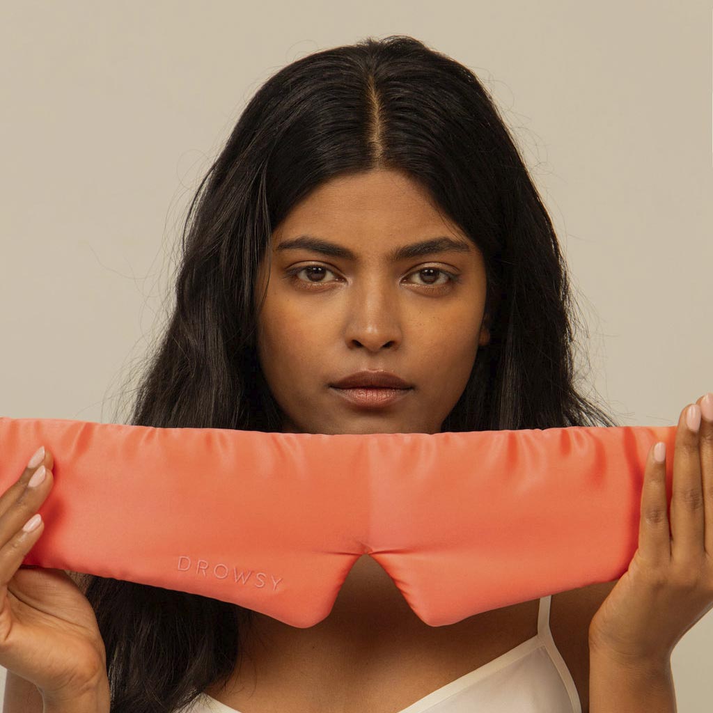 A woman with long dark hair holds a “Summer Coral” silk eye mask in front of her chest, looking at the camera against a plain beige background.
