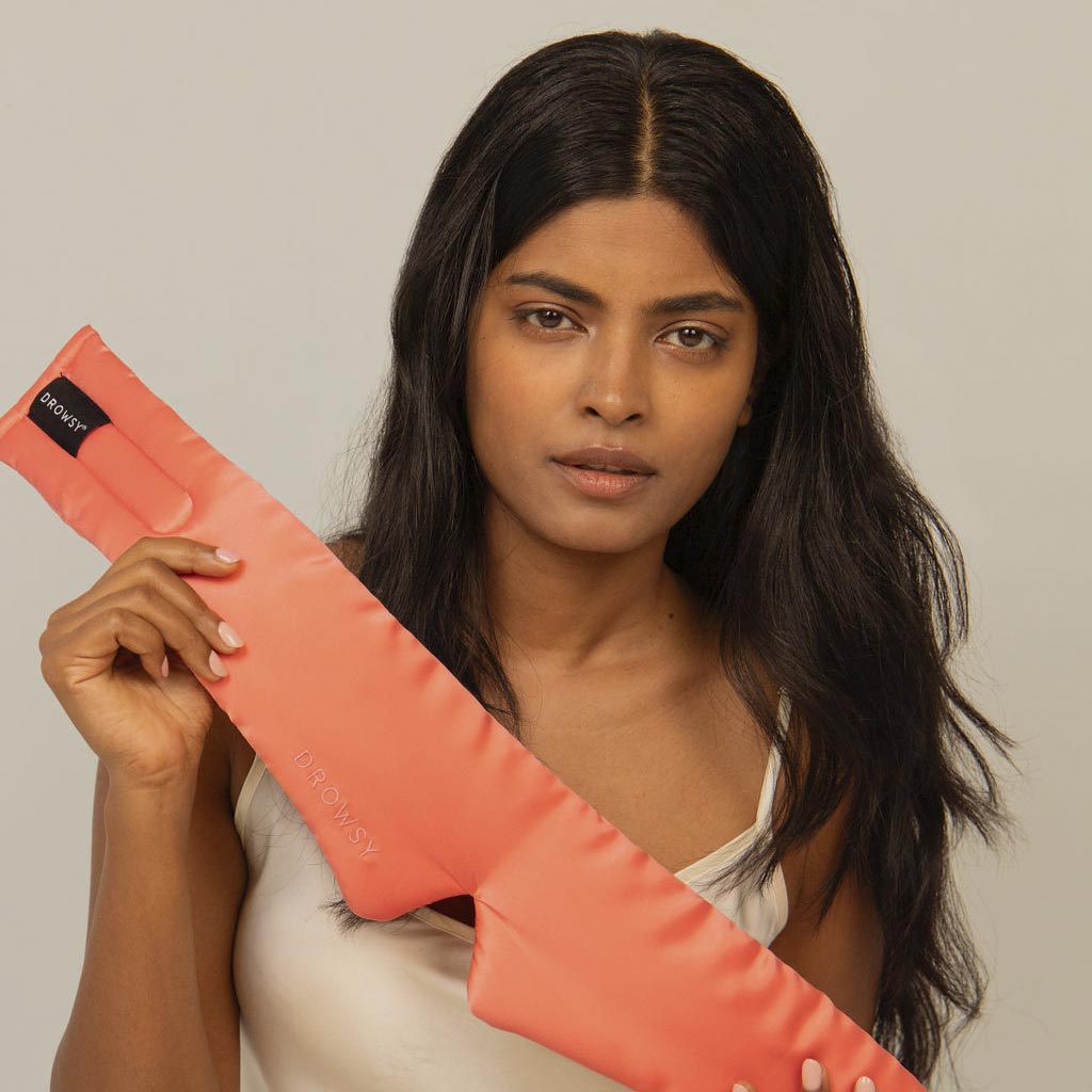 A woman with long dark hair and a neutral expression holds the Summer Coral silk eye mask, wearing a white tank top against a plain background—perfect for beauty sleep.