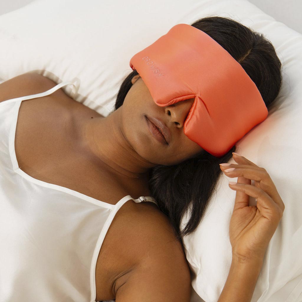 A woman relaxes on a white pillow, wearing the Summer Coral large orange silk eye mask and a white satin camisole. The mask covers her eyes and nose as she enjoys restful beauty sleep, one hand resting near her face.