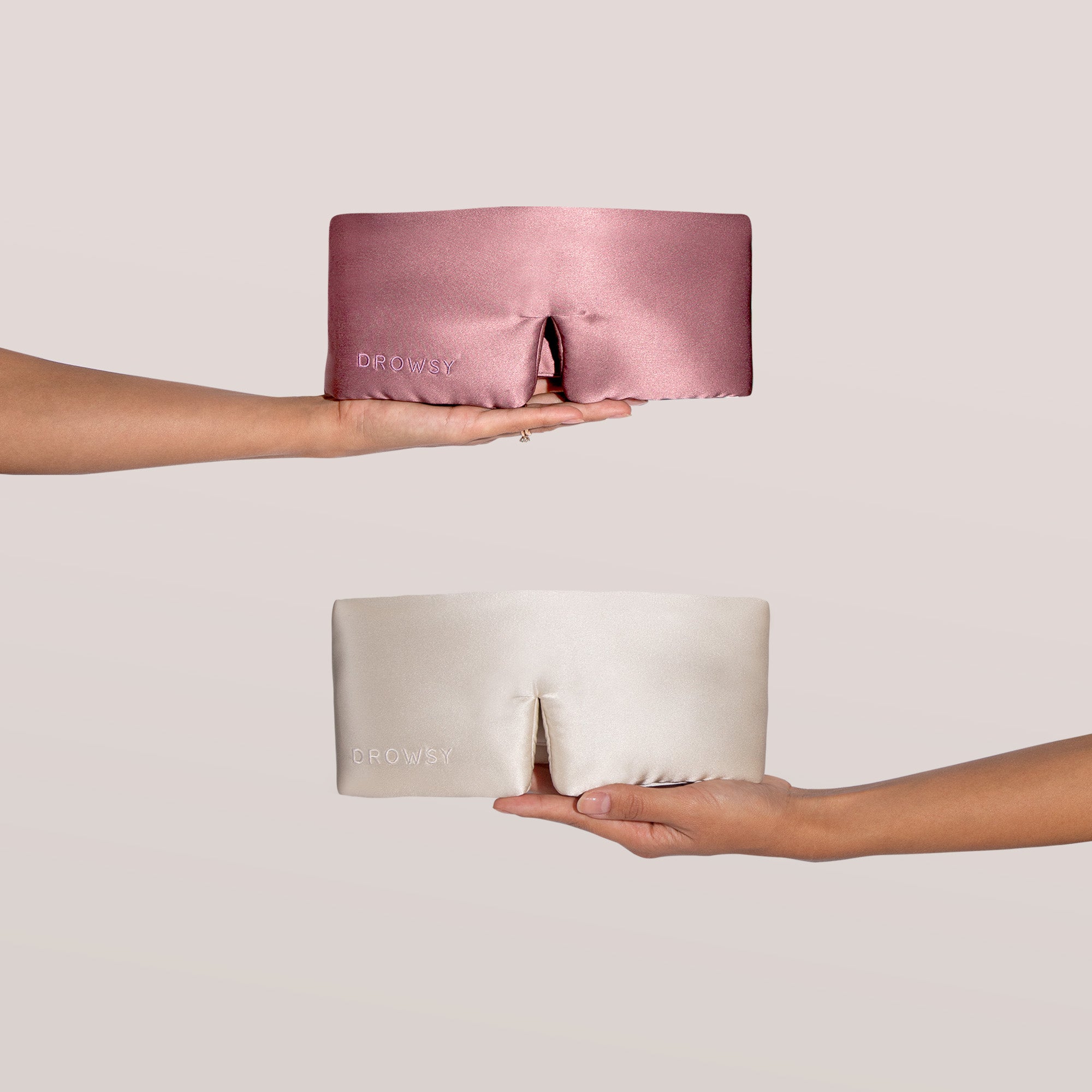 Two hands each hold a silk sleep mask with a nose cutout. The top mask is pink and the bottom is cream, both with DROWSY embroidered on the front. The background is light and neutral.