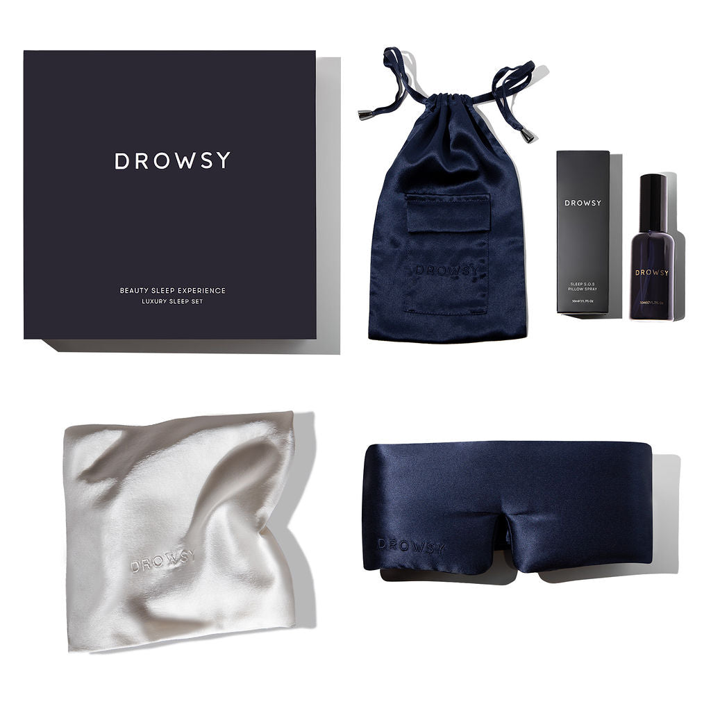 A luxury sleep set includes a dark gift box, a navy blue satin pouch, a black bottle with box, a white silk pillowcase, and a black silk eye mask, all labeled Drowsy on a white background.