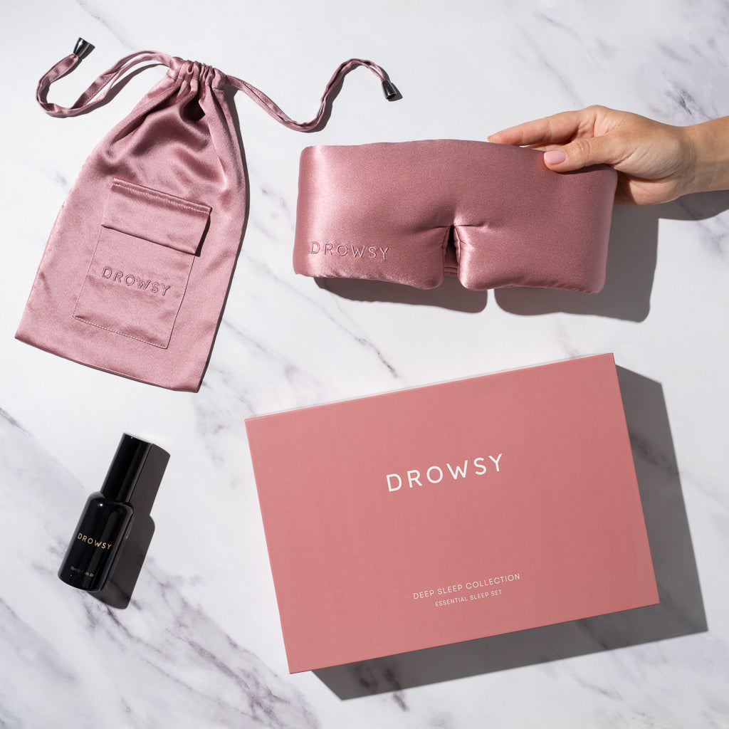 A hand holds a luxury Damask Rose - Deep Sleep Collection silk sleep mask with a matching pouch, black deep sleep pillow spray bottle, and pink box—all on white marble. Perfect sleep essentials.