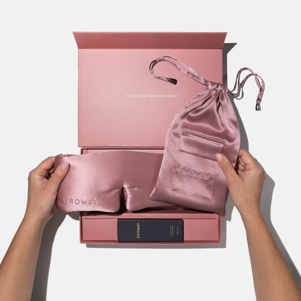 A person holds the Damask Rose - Deep Sleep Collection luxury silk sleep mask with matching pink carry pouch above an open pink gift box with “You deserve glorious sleep” inside the lid and a small product box nestled within.