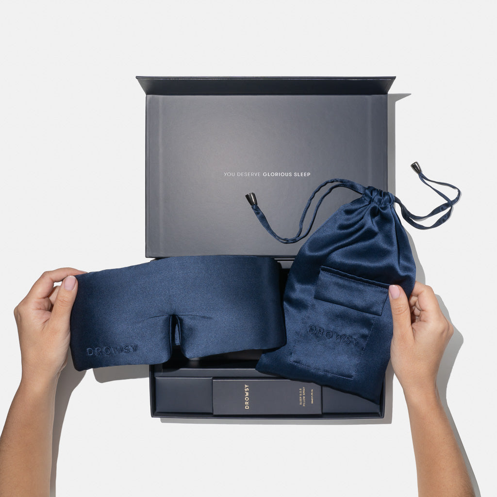 A person holds a navy blue silk sleep mask and matching pouch from the Midnight Blue - Deep Sleep Collection in front of an open gift box reading “YOU DESERVE GLORIOUS SLEEP,” with luxury sleep essentials neatly arranged inside.