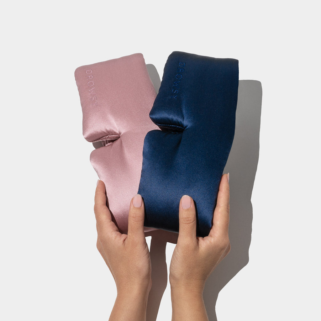 Two hands hold the Lovers Pack sleep masks, one pink and one navy blue, against a plain white background. Both satin masks feature a smooth, shiny texture with a subtle embossed logo on the fabric.