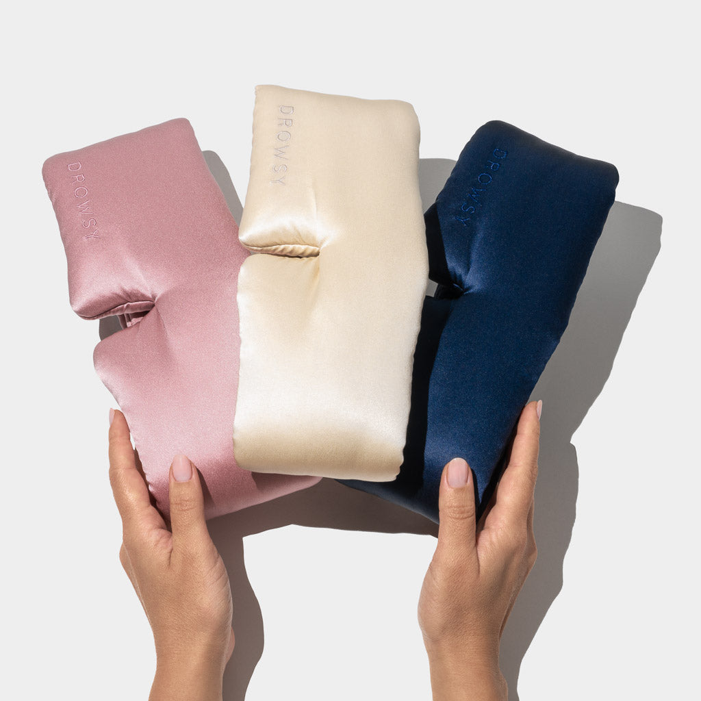 Two hands hold the Besties Pack—three silky, cushion-like sleep masks in pink, cream, and navy blue—against a plain light background.