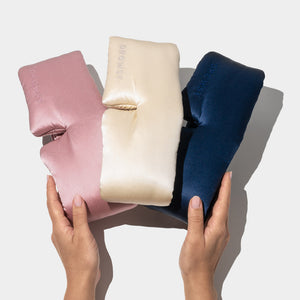 Two hands hold the Besties Pack—three silky, cushion-like sleep masks in pink, cream, and navy blue—against a plain light background.