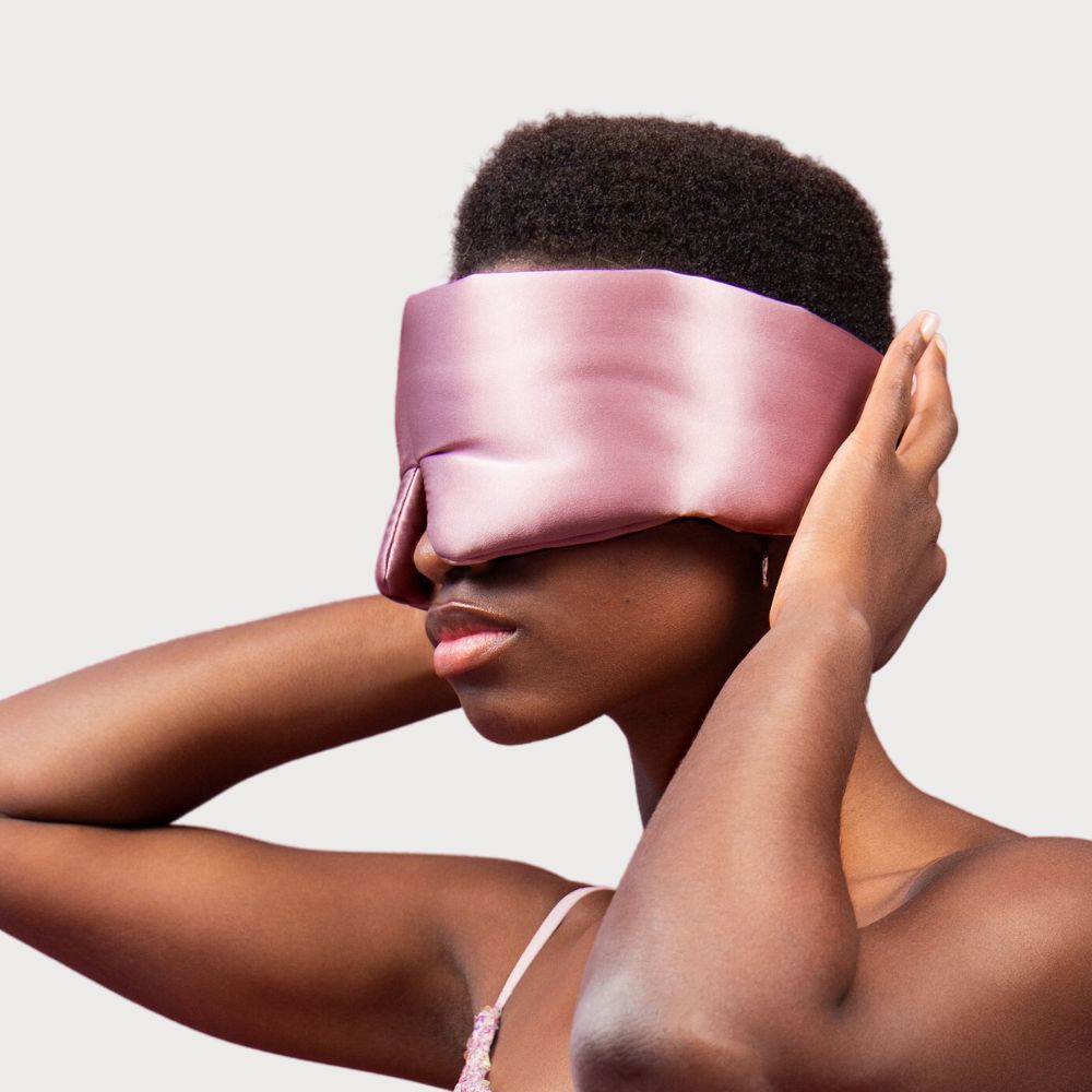 A person with short hair wears a shiny, oversized pink sleep mask, covering their eyes and part of their face, while holding it in place with both hands against a plain light background.