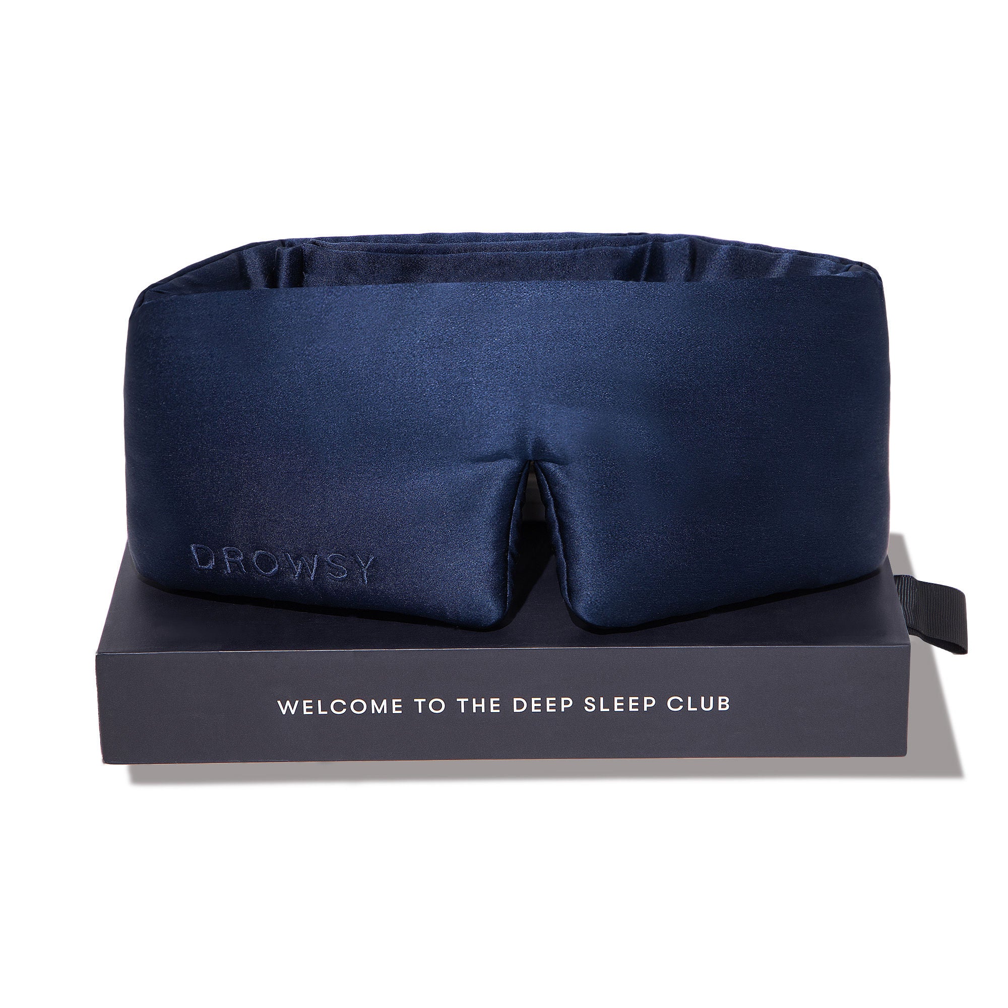 A navy blue satin sleep mask with the word DROWSY embossed on the side, displayed on a black box that says WELCOME TO THE DEEP SLEEP CLUB in white letters.