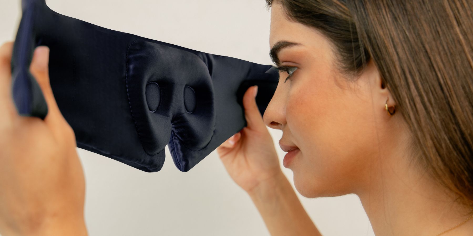 A woman holds a navy blue, padded sleep mask with raised eye cups, preparing to put it on her face. She has straight brown hair and wears a small hoop earring. The background is plain and light-colored.