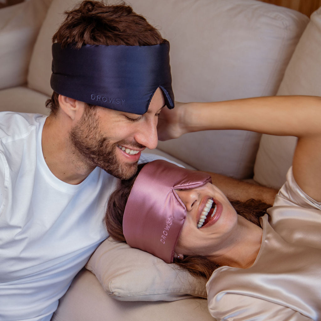 A smiling man and woman relax on a sofa, each wearing a silk sleep mask—his is navy blue and hers is pink. The woman is lying down, laughing, while the man leans in close, both appearing happy and comfortable.