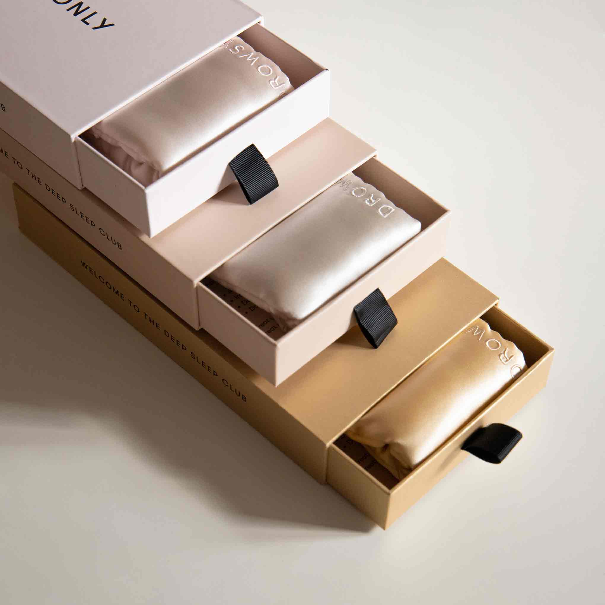 Three rectangular boxes—two cream-colored, one tan—are stacked and partially open, each revealing a satin pouch inside. The pouches have a black pull tab and white embroidered text reading DROWSY.