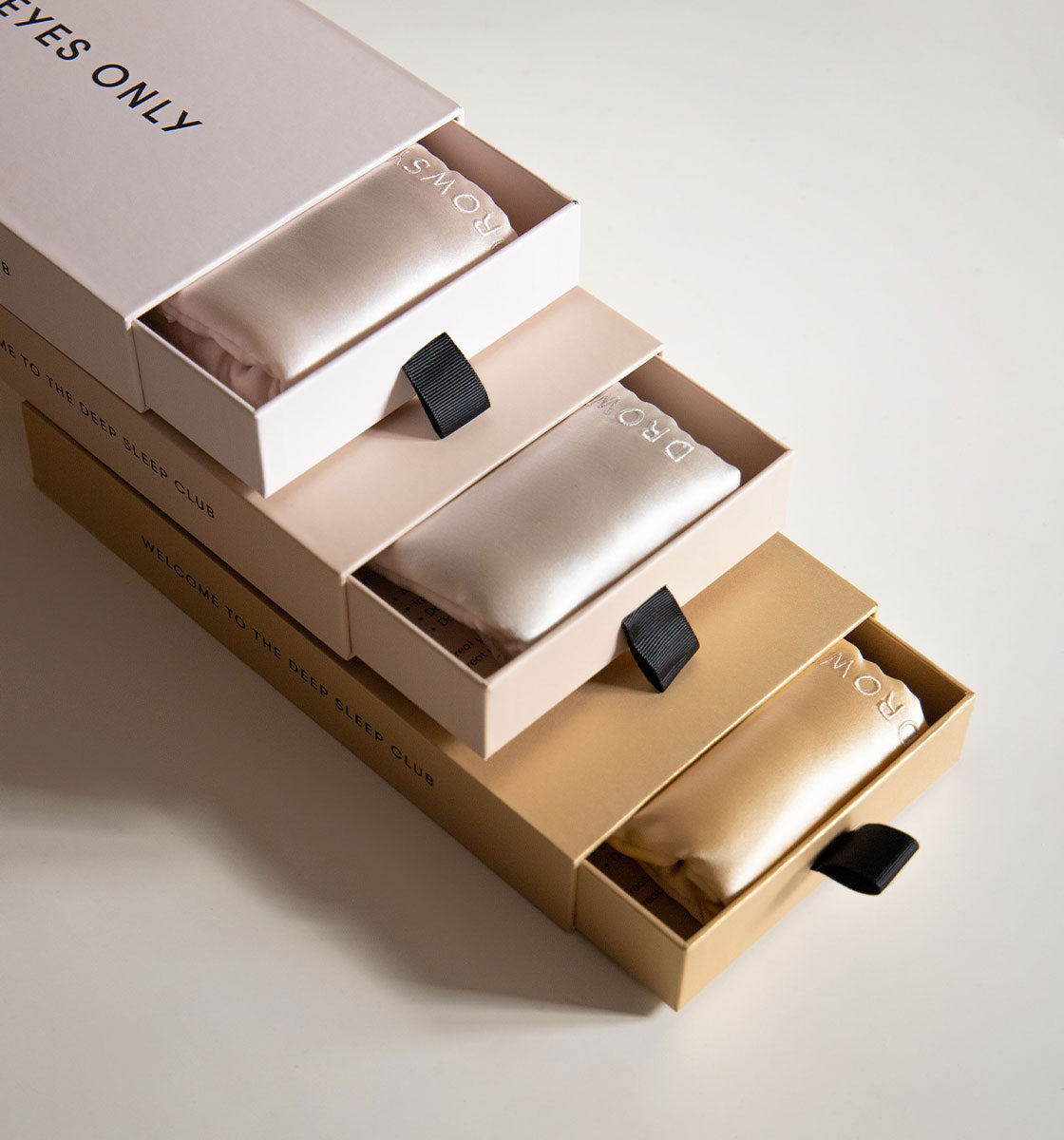 Three satin sleep masks in pink, cream, and gold rest in matching sliding boxes, each with a black pull tab and subtle branding, arranged on a light surface.