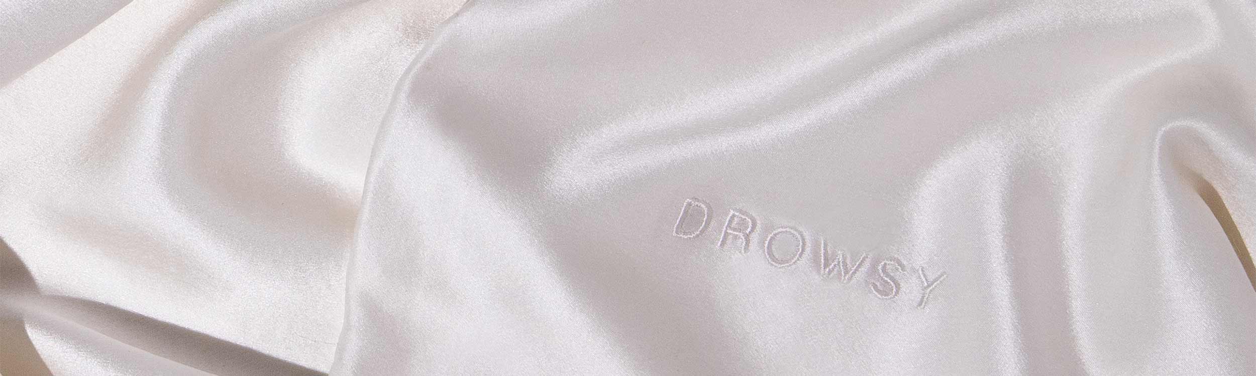 A close-up of smooth, white satin fabric with the word DROWSY embroidered in matching white thread.