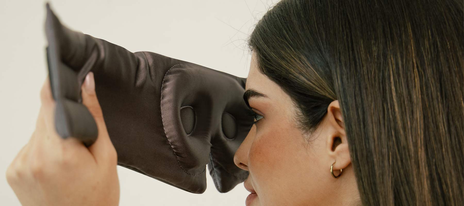 A woman with straight brown hair holds a large, padded black sleep mask with eye cutouts close to her face, preparing to put it on.
