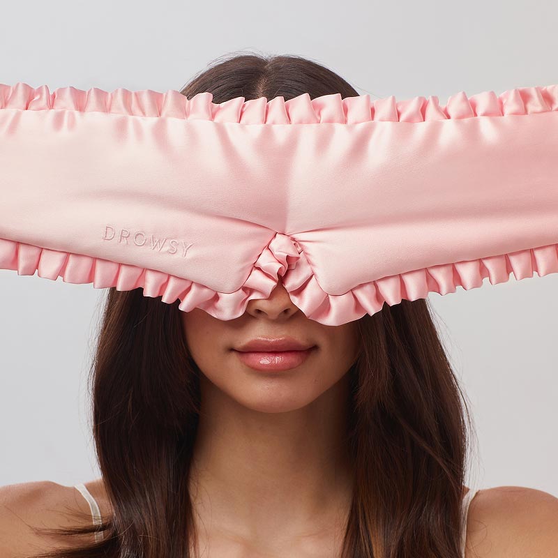A woman with long brown hair wears drowsysleepco’s Manhattan Experience Bundle—a large, ruffled pink satin sleep mask embroidered with “DROWSY”—covering her eyes and eyebrows against a plain light background.
