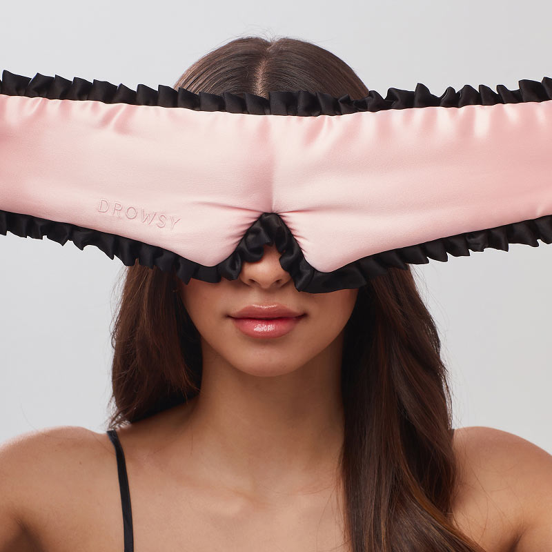 A woman with long brown hair wears drowsysleepco's Manhattan Experience Bundle—a large, light pink sleep mask with black ruffled edges and "DROWSY" embossed—covering her eyes. She has a neutral expression and wears a black strap top.
