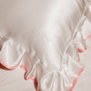The drowsysleepco Sleep and the City - Pearl & Pink Pillowcase, featuring "DROWSY" embroidered in the center and pink-edged ruffled trim, rests on a bed.