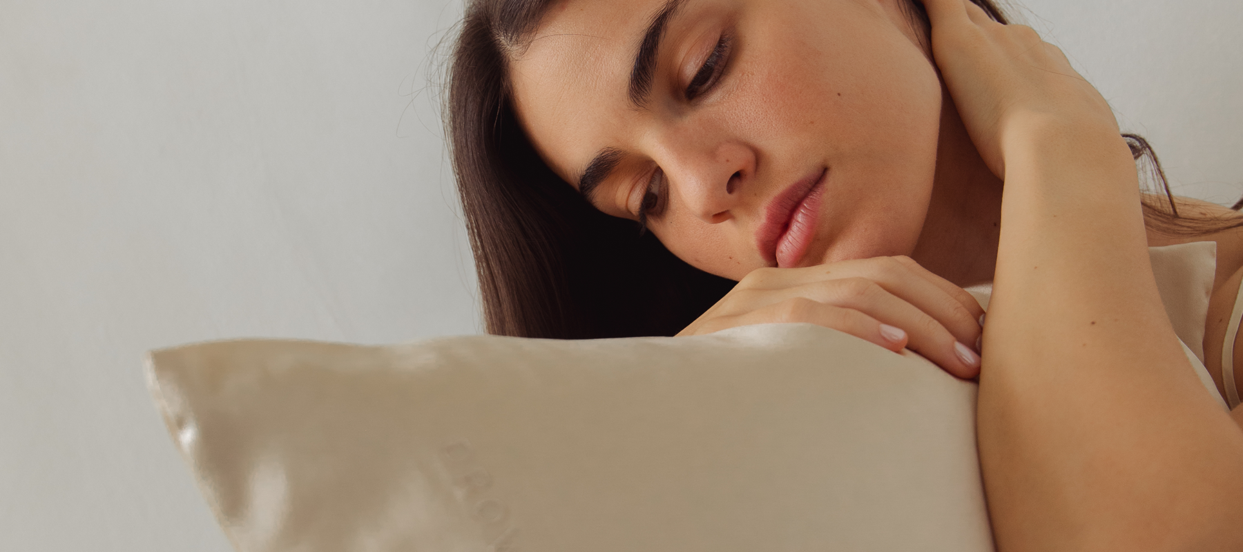 A woman with long dark hair rests her head and hand on a light-colored silk pillow, looking down thoughtfully with a serene expression against a plain background.