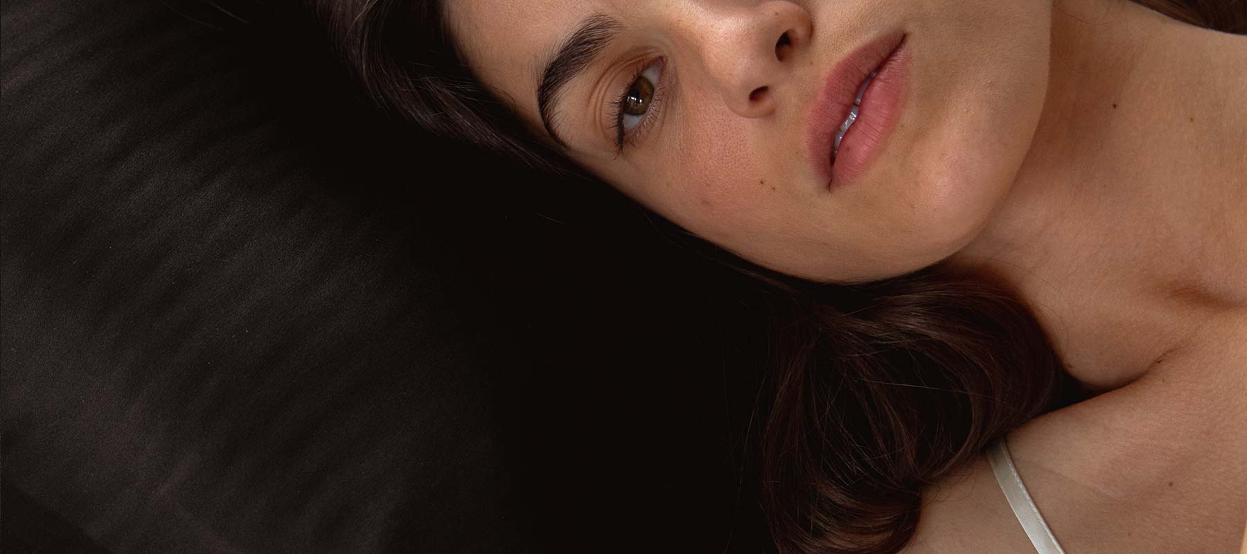 A woman with dark hair lies on a black surface, gazing softly at the camera. She wears a white strap, and only part of her face and shoulder are visible in a close-up shot.