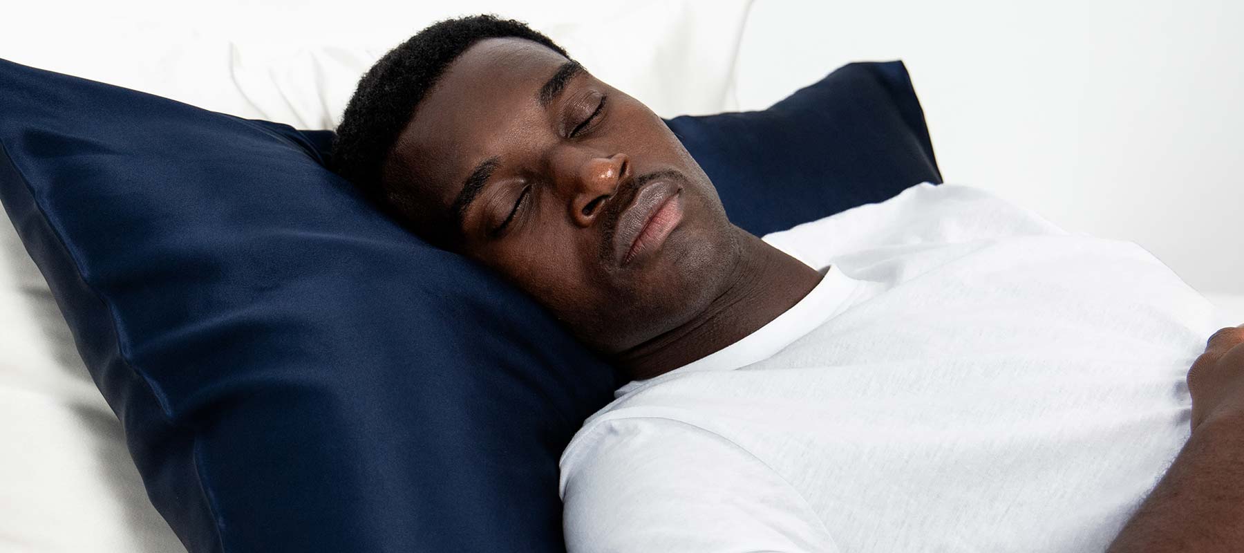 A man wearing a white T-shirt is lying on his back with his eyes closed, resting on a dark blue pillow in a bright, minimalistic setting.