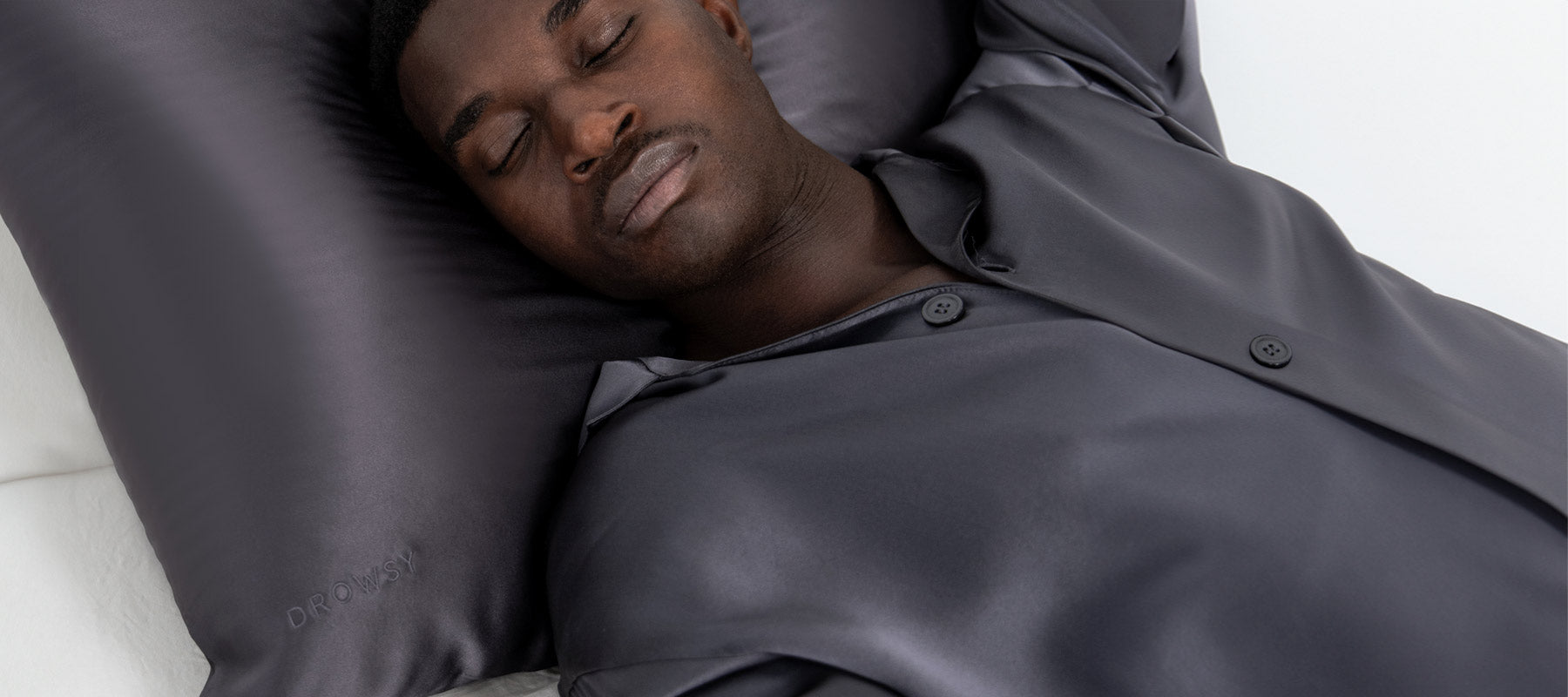 A man wearing dark satin pajamas sleeps on his back with his arms above his head, resting on a matching satin pillow.