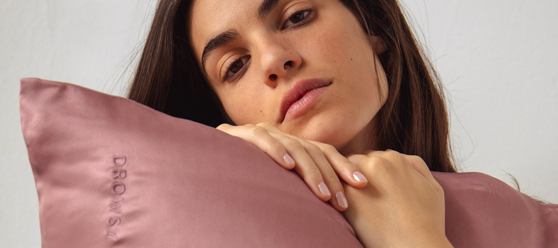 A woman with long brown hair rests her head and hands on a mauve satin pillow embroidered with the word DROWSY, looking calmly at the camera against a plain light background.