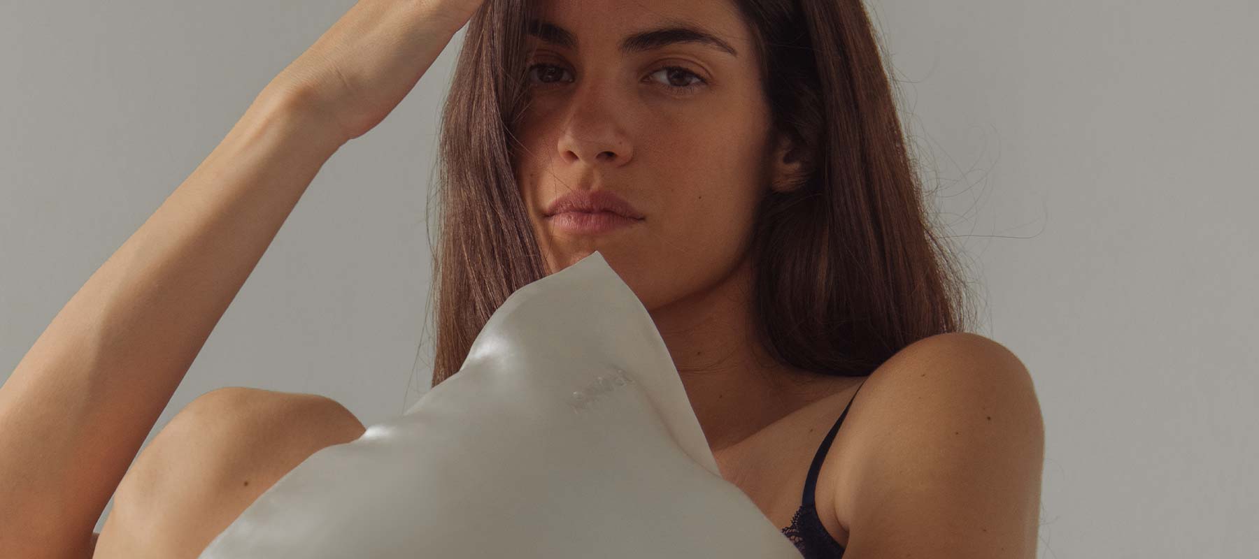 A woman with long brown hair holds a white pillow, looking directly at the camera with a neutral expression. She wears a dark top, and soft natural light illuminates her face against a plain background.