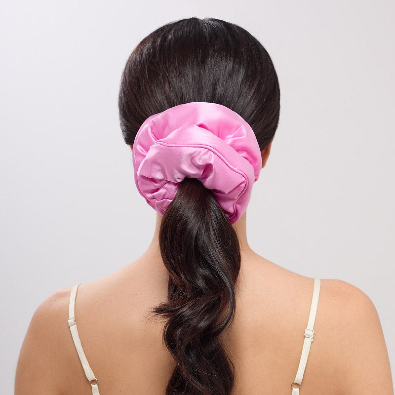 A woman with long, dark hair is seen from behind, wearing a spaghetti strap top and the Hotel Paradiso - Heatwave Padded Scrunchie in hot pink, securing her low ponytail against a plain, light-colored background.