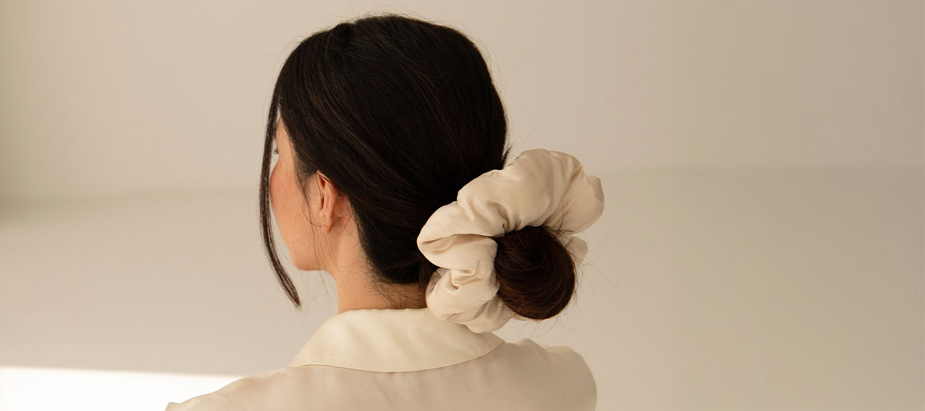 A woman with long dark hair in a low bun, secured with a large beige scrunchie, is seen from behind against a plain, light background. She is wearing a cream-colored top.
