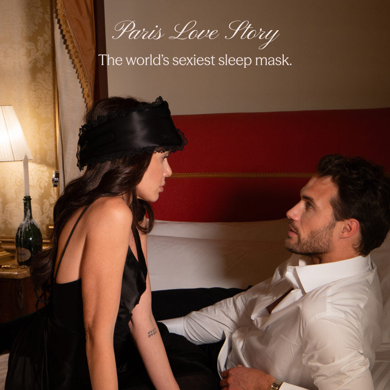 A woman wearing the Paris Love Story - Black Jade Sleep Mask by drowsysleepco sits on a bed facing a man in a white shirt. Soft lighting and elegant decor set a romantic mood. Text: The world’s sexiest sleep mask.