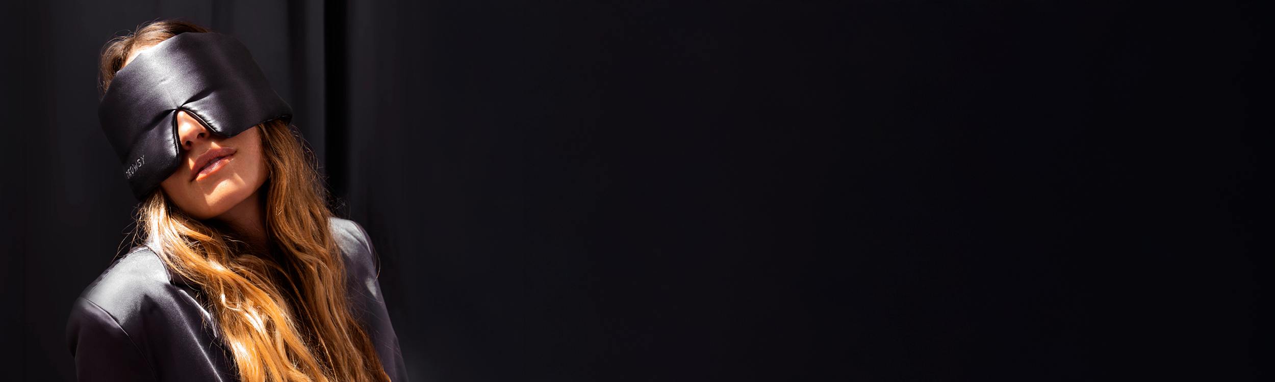 A person with long hair wearing a dark eye mask, tilting their head slightly back, set against a dark background with light highlighting the left side of their face and hair.