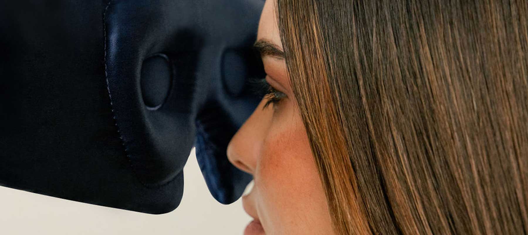 A woman with straight brown hair uses a black padded eye mask or face cushion, pressing her face gently against its surface.